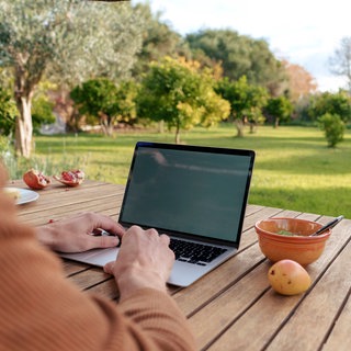 Digitaler Nomade arbeitet im Garten am Laptop: Manche Kommunen reagieren mit kreativen Strategien auf den Leerstand – mit Probewohnen, Zuschüssen für Baugrundstücke, gezieltem Standortmarketing oder der Errichtung digitaler Nomadendörfer.