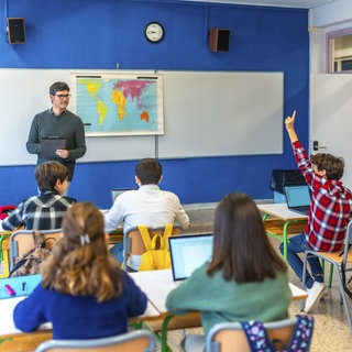 Eine Gruppe Jugendlicher hebt die Hand und folgt dem Erdkundeunterricht im Klassenzimmer mit Lehrer und Laptops.