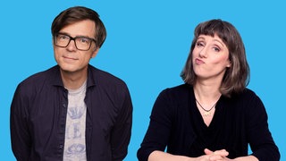 Ein Mann und eine Frau sitzen zusammen an einem Tisch und schauen in die Kamera. Die Host des Podcasts "Fakt ab! Eine Woche Wissenschaft" und ihr Gast Ralph Caspers