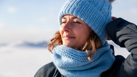 Frau mit Mütze reckt in Winterlandschaft das Gesicht in die Sonne:  Licht beeinflusst fast alle Bereiche unseres Lebens. Die Interaktion von Licht und Farbe prägt unmittelbar unsere Wahrnehmung. 