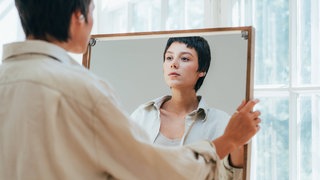 Eine Frau hält sich selbst einen großen Spiegel vor: Narzissmus zeigt sich bei Frauen oft als emotionale Abhängigkeit. Minderwertigkeitskomplexe sind die verletzliche Seite ihres narzisstischen Selbstbilds und können zu toxischem Verhalten führen.