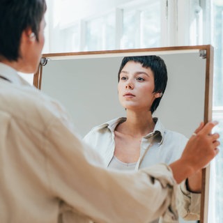 Eine Frau hält sich selbst einen großen Spiegel vor: Narzissmus zeigt sich bei Frauen oft als emotionale Abhängigkeit. Minderwertigkeitskomplexe sind die verletzliche Seite ihres narzisstischen Selbstbilds und können zu toxischem Verhalten führen.