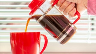 Frischer Kaffee wird aus French Press in eine rote Tasse gegossen: Dass Kaffee oder Schwarztee dem Körper Wasser entzieht, hat man mal geglaubt. Das ist aber längst überholt.
