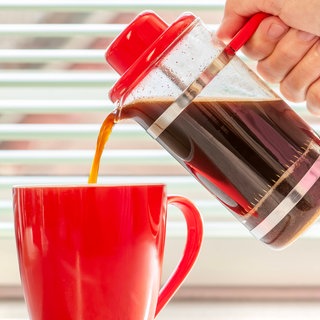 Frischer Kaffee wird aus French Press in eine rote Tasse gegossen: Dass Kaffee oder Schwarztee dem Körper Wasser entzieht, hat man mal geglaubt. Das ist aber längst überholt.