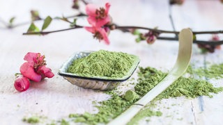 Matcha-Pulver, rosa Blüten und ein Matcha-Löffel aus Bambus: Kurzfristig steigert grüner Tee die Konzentrationsfähigkeit, oft auch die Stimmung, das Koffein kann leicht euphorisierend wirken. Krankheiten beugt er allein nicht vor. 