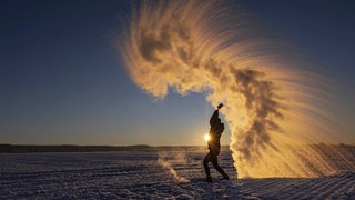 Eine Frau nutzt den Mpemba-Effekt: Sie wirft heißes Wasser in die Luft, das spektakulär zu Eiskristallen gefriert. Warum gefriert warmes Wasser schneller als kaltes?
