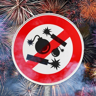 Schild mit durchgestrichenen Böllern und Kugelbomben, dahinter Feuerwerk: Ein Feuerwerk kann ein Kunstwerk sein. Dafür ist eine Ausbildung nötig. An Silvester wird an vielen Orten einfach nur geböllert und geknallt. Die Polizei muss entscheiden, wo sie kontrolliert.