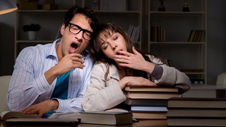 Ein Mann und eine Frau sitzen gähnend über einem Stapel Bücher: Warum ist gähnen ansteckend?