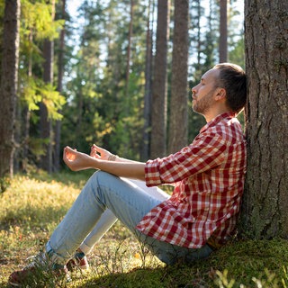 Ein Mann sitzt an einem sonnigen Tag mit geschlossenen Augen unter einer Kiefer im Wald und meditiert: Die Hälfte der Deutschen fühlt sich erschöpft. Doch Erholung braucht mehr als nur Ruhe. Sie muss zu unseren Bedürfnissen passen. 