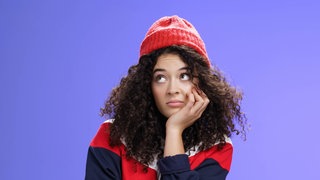 Junge Frau mit roter Mütze und lockigem Haar schaut gelangweilt: Langeweile ist ein den meisten Menschen vertrautes Gefühl, in der Wissenschaft aber noch nicht besonders gut erforscht. Wann ist Langeweile gut, wann schadet sie?