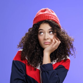 Junge Frau mit roter Mütze und lockigem Haar schaut gelangweilt: Langeweile ist ein den meisten Menschen vertrautes Gefühl, in der Wissenschaft aber noch nicht besonders gut erforscht. Wann ist Langeweile gut, wann schadet sie?