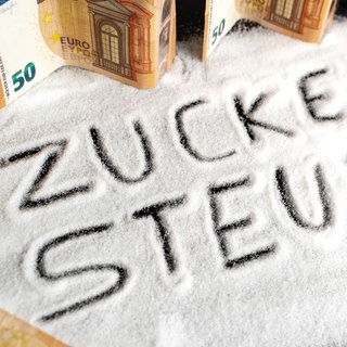Euro-Geldscheine liegen auf Zucker, in den das Wort Zuckersteuer geschrieben ist.