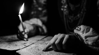 Frau liest mit brennender Kerze in der Hand in einem alten Buch: Sagt man mir brennt etwas auf den Nägeln oder unter den Nägeln? Und woher kommt die Redensart?