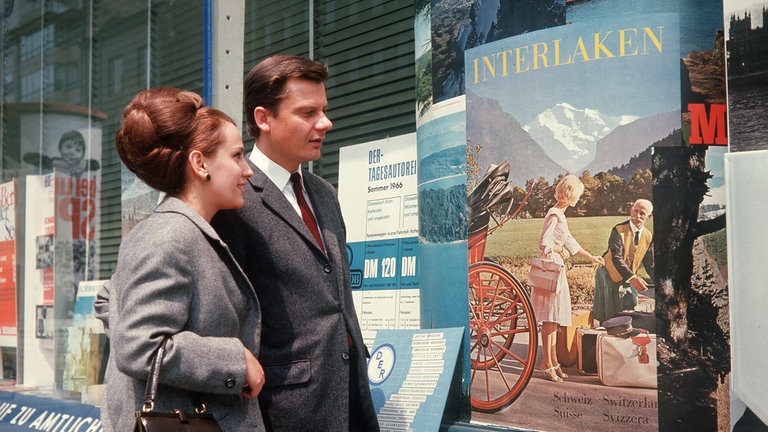Junges Paar steht in den 1960ern vor einem Reisebüro und betrachtet Plakatwerbung fürs schweizerische Interlaken: Nach Ende des Zweiten Weltkriegs erfuhr die Bundesrepublik einen enormen wirtschaftlichen Aufschwung. Reisen, Möbel, Autos waren begehrt.