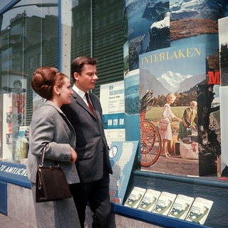 Junges Paar steht in den 1960ern vor einem Reisebüro und betrachtet Plakatwerbung fürs schweizerische Interlaken: Nach Ende des Zweiten Weltkriegs erfuhr die Bundesrepublik einen enormen wirtschaftlichen Aufschwung. Reisen, Möbel, Autos waren begehrt.