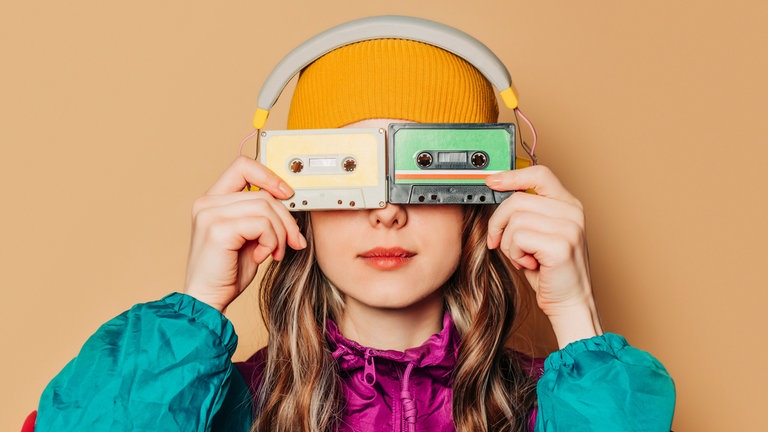 Junge Frau im Ballonseiden-Sportanzug hält sich Audiokassetten vor die Augen: Musik aus den 1980ern, Mode aus den 1990ern: Manche junge Menschen sehnen sich zurück in eine Zeit, die sie selbst nicht erlebt haben. 