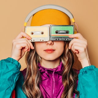 Junge Frau im Ballonseiden-Sportanzug hält sich Audiokassetten vor die Augen: Musik aus den 1980ern, Mode aus den 1990ern: Manche junge Menschen sehnen sich zurück in eine Zeit, die sie selbst nicht erlebt haben. 