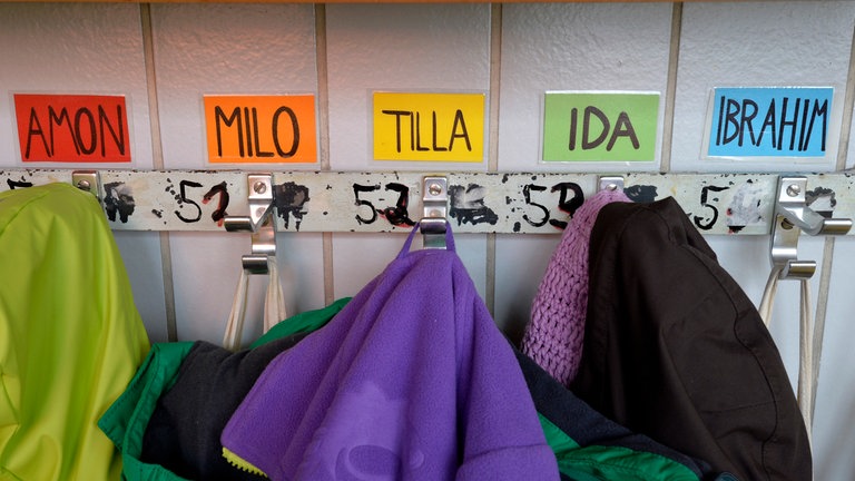 Kleiderhaken mit Namen von Kindern verschiedener Nationalitäten: Amon, Milo, Tilla, Ida, Ibrahim. – Namen geben Hinweise auf Herkunft und die Identität. Sie können aber auch zur Last werden, denn oft sind sie mit Vorurteilen belegt.