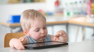 Ein kleiner Junge tippt auf einem Tablet: Ob Tablet oder Smartphone: Kleinkinder kommen etwa mit eins damit erstmals in Kontakt. Fachleute warnen und fordern: "Bildschirmfrei bis drei". Aber die Studienlage ist uneindeutig. Es kommt auf die Art der Nutzung an.