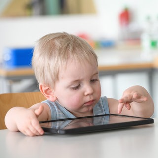 Ein kleiner Junge tippt auf einem Tablet: Ob Tablet oder Smartphone: Kleinkinder kommen etwa mit eins damit erstmals in Kontakt. Fachleute warnen und fordern: "Bildschirmfrei bis drei". Aber die Studienlage ist uneindeutig. Es kommt auf die Art der Nutzung an.