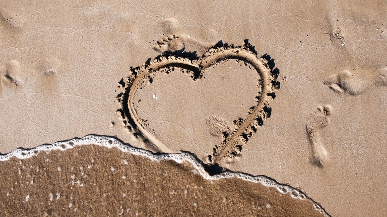 Herz, in Sandstrand gezeichnet, wird von Meeresflut weggespült: Trennungen kommen selten plötzlich. Meist ist vorher schon die Zufriedenheit in der Beziehung über Jahre zurückgegangen und Nähe in Distanz umgeschlagen. 