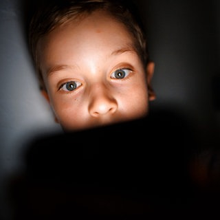 Symbolfoto: Gestellte Aufnahme zum Thema Mediennutzung bei Kindern. Ein Kind schaut im Dunkeln auf das Display eines Smartphones.