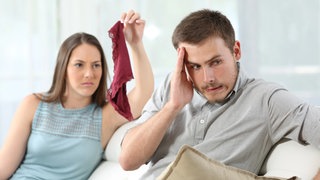 Erwischt! Wütende junge Frau präsentiert ihrem beschämten Partner fremde rote Damenunterwäsche: Treue gilt als Basis für eine erfüllte Liebesbeziehung. Ein Seitensprung ist daher die häufigste Ursache für Konflikte und Trennungen.