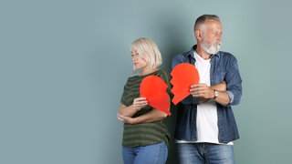 Grauhaariges Paar, Mann und Frau, stehen nebeneinernder mit abgewandten Gesichtern und halten ein zerrissens rotes Papierherz: Immer häufiger trennen sich Paare nach langer Ehe. Warum wächst die Zahl der "grauen Scheidungen"?