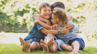 Drei kleine Geschwister umarmen sich lachend auf einer Wiese: Geschwister gehören zu den ersten und oft einflussreichsten Beziehungen unseres Lebens. Die Rollen, die wir miteinander aushandeln, können uns auch weit übers Familienleben hinaus prägen. 