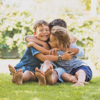 Drei kleine Geschwister umarmen sich lachend auf einer Wiese: Geschwister gehören zu den ersten und oft einflussreichsten Beziehungen unseres Lebens. Die Rollen, die wir miteinander aushandeln, können uns auch weit übers Familienleben hinaus prägen. 