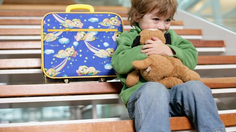 Kind mit kleinem Koffer sitzt auf einer Treppe und drückt sein Gesicht an einen Teddybär: Jedes Jahr werden in Deutschland Tausende Kinder vom Jugendamt in Obhut genommen. Ist das für die betroffene Familien wirklich eine Hilfe?