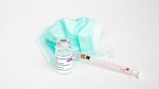 Vor einem weissen Hintergrund ist eine Ampulle mit dem COVID-19-Vektor-Impfstoff von AstraZeneca zu sehen, daneben liegt ein Mundschutz und eine Impfspritze für die Impfung.