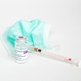Vor einem weissen Hintergrund ist eine Ampulle mit dem COVID-19-Vektor-Impfstoff von AstraZeneca zu sehen, daneben liegt ein Mundschutz und eine Impfspritze für die Impfung.