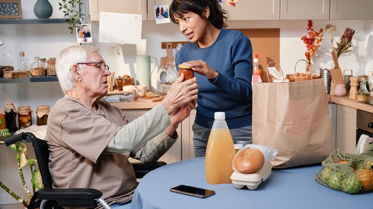 Ehrenamtliche hat für älteren Nachbarn im Rollstuhl Lebensmittel eingekauft und packt sie am Tisch aus: Nachbarschaftshilfe ist eins der Konzepte, damit Senioren möglichst lange zu Hause leben können.