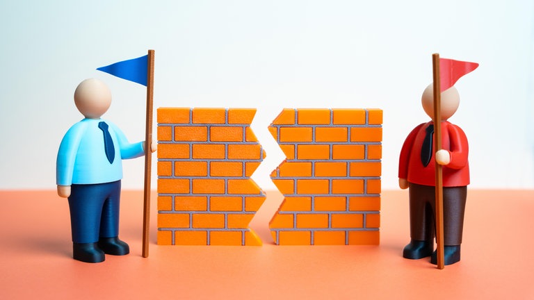 Zwei Rivalen mit je einem blauen und einem roten Fähnchen und ein Riss in einer Mauer symbolisieren Konflikte: Nicht nur Argumente beeinflussen unsere politische Meinungsbildung. Auch Emotionen und Bedürfnisse.