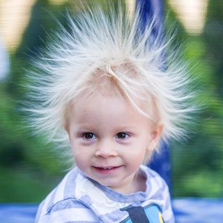 Kleiner blonder Junge. Seine Haare sind statisch geladen und stehen rund um seinen Kopf in die Höhe: Warum sind wir manchmal elektrisch aufgeladen)