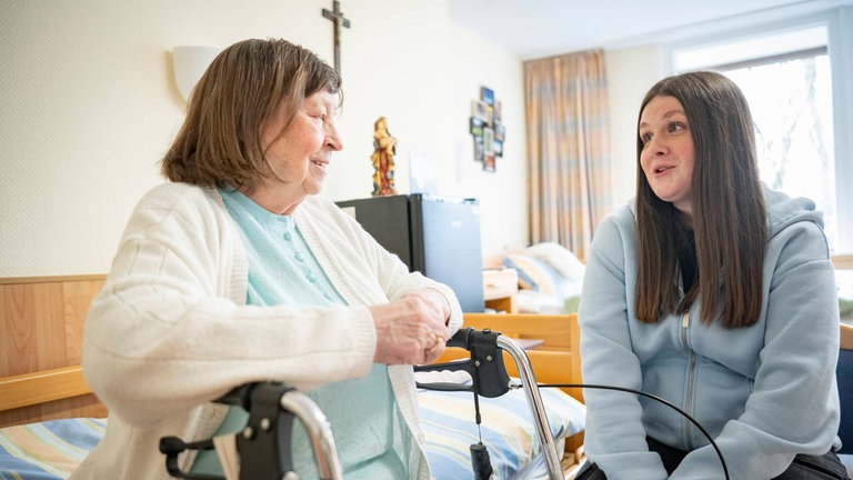 Junge Frau im Bundesfreiwilligendienst unterhält sich mit einer älteren Frau im Seniorenheim: Die Politik fordert ein verpflichtendes Gesellschaftsjahr für alle. Okay so – oder sollte das Engagement freiwillig bleiben? (Symbolbild)
