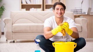 Mann sitzt lachend und entspannt im Schneidersitz hinter einem Putzeimer: Viele Männer betonen, wie wichtig ihnen Gleichberechtigung in der Partnerschaft ist – doch die Realität sieht anders aus. 