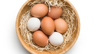 Schale mit weißen und braunen Eiern auf beigefarbenem Ostergras: Wovon hängt die Farbe der Eier ab?