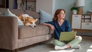 Junge Frau sitzt im Schneidersitz mit Laptop auf dem Boden: Sitzt man längere Zeit im Schneidersitz, kann es passieren, dass die Füße oder Beine einschlafen. Bei manchen Leuten passiert das auch, wenn sie zu enge Schuhe tragen.