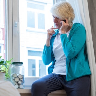 Eine ältere Frau sitzt in einer Wohnung am Fenster und telefoniert (gestellte Szene).