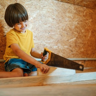 Symbolbild: Lächelndes Kind im gelben Hemd sägt Holz bei Hausrenovierung 