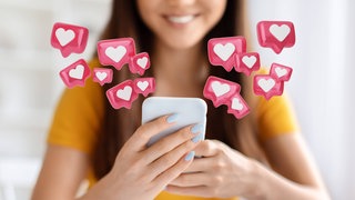 Junge Frau schaut glücklich lächelnd auf ihr Smartphone, aus dem Herzen fliegen: Bewusstes Dopamintraining mit genügend Schlaf, Pausen und echten Zielen statt Likes und Push-Nachrichten stabilisiert uns im digitalen Alltag.