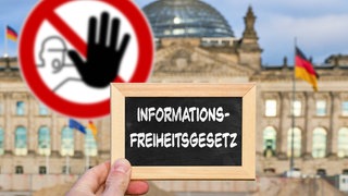 Hand mit Tafel vor dem Reichstagsgebäude, darauf der Schriftzug "Informationsfreiheitsgesetz". Im Hintergrund ein Warnschild mit erhobener Hand: Um die Informationsfreiheit steht es in Deutschland nicht besonders gut