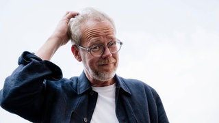 Symbolfoto: Porträt einer bärtigen Person mit Brille, die sich im Freien am Kopf kratzt. 