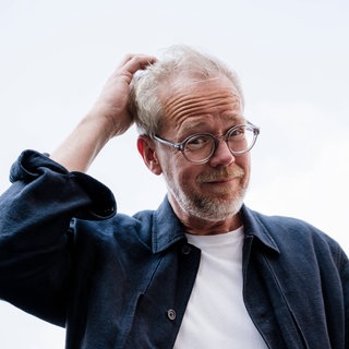 Symbolfoto: Porträt einer bärtigen Person mit Brille, die sich im Freien am Kopf kratzt. 