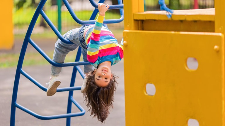 Kleines Mädchen hängt kopfüber an einem Klettergerüst: Bei Spielplätzen prallen Interessen aufeinander. Kinder wollen spielen, Eltern Sicherheit, Kommunen DIN-Normen einhalten und die sinkenden Budgets abfedern. 