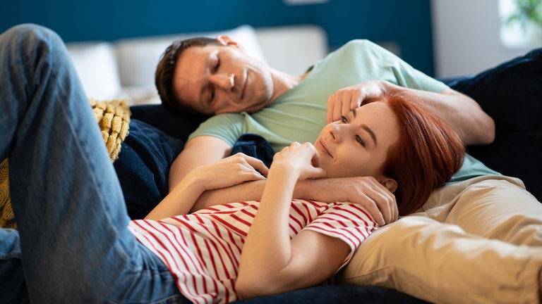 Junges Paar kuschelt auf dem Sofa: Bindungshormon, Treue- oder Orgasmushormon: Oxytocin werden Superkräfte nachgesagt. Zur Superkraft für unser Wohlbefinden wird es aber nur, wenn wir mit einem anderen Menschen zugewandt in Beziehung stehen. 