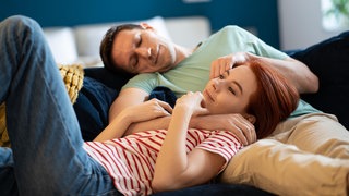 Junges Paar kuschelt auf dem Sofa: Bindungshormon, Treue- oder Orgasmushormon: Oxytocin werden Superkräfte nachgesagt. Zur Superkraft für unser Wohlbefinden wird es aber nur, wenn wir mit einem anderen Menschen zugewandt in Beziehung stehen. 
