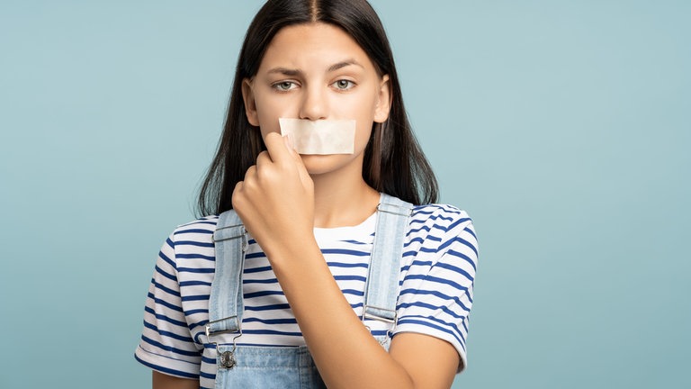Junge Frau reißt sich Pflaster vom Mund: Tabus sind oft negativ behaftet. Veraltete Gebote, denen wir uns unterordnen. Sie ziehen aber auch Grenzen, die nicht verhandelbar sind. Der Schutz von Kindern etwa.